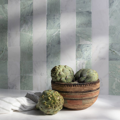 A woven basket filled with artichokes sits on a white surface against a wall of vertical marble tiles in pale green and white. A folded white cloth is partly visible on the left.