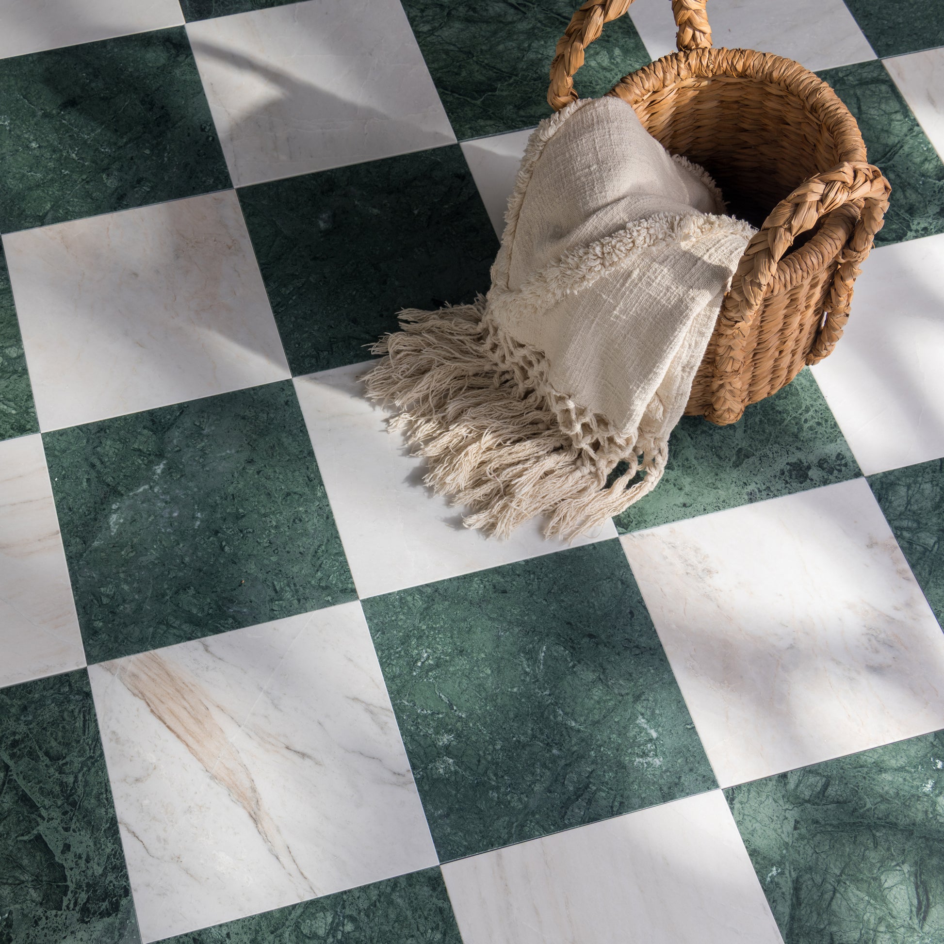 A styled floor scene with a woven basket holding a cream fringed throw blanket, placed on a green-and-white checkered marble floor.