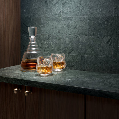 A clear glass whiskey decanter and two matching lowball glasses filled with amber liquor, set on a dark stone countertop against a dark green marble tile backsplash.