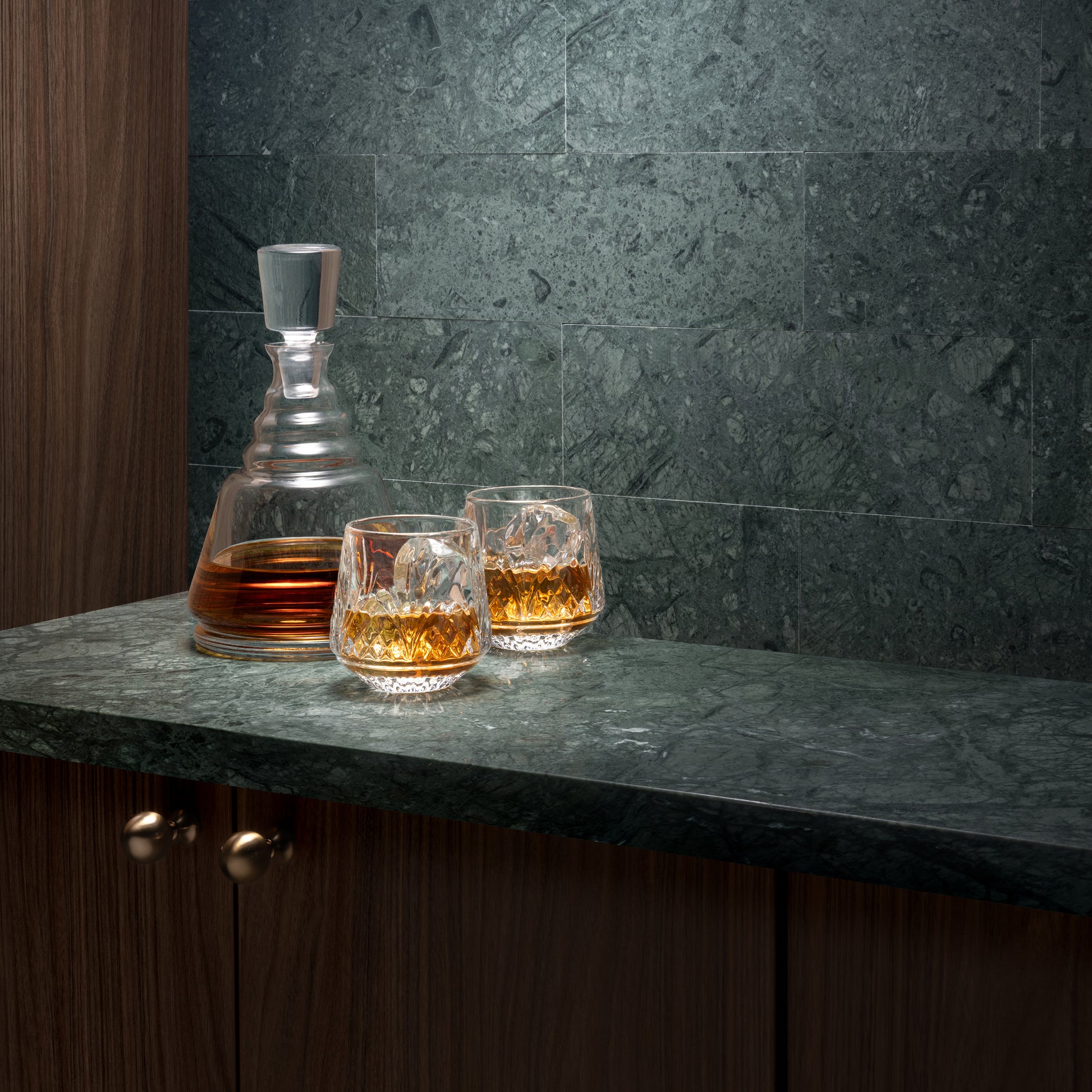 A clear glass whiskey decanter and two matching lowball glasses filled with amber liquor, set on a dark stone countertop against a dark green marble tile backsplash.