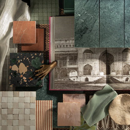 A styled flat-lay of home decor and material samples, including marble and terrazzo tiles, fabric swatches, wallpaper, a book with an architectural illustration, and a small metallic hand sculpture on a dark green textured surface.