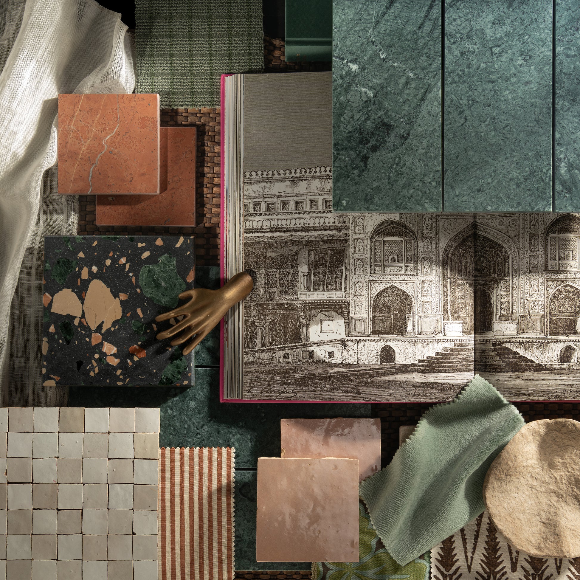 A styled flat-lay of home decor and material samples, including marble and terrazzo tiles, fabric swatches, wallpaper, a book with an architectural illustration, and a small metallic hand sculpture on a dark green textured surface.