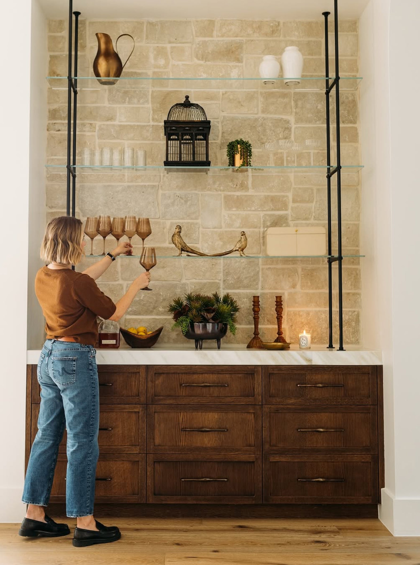 Person arranging items on a shelf in a kitchen with a stone wall and wooden cabinets.