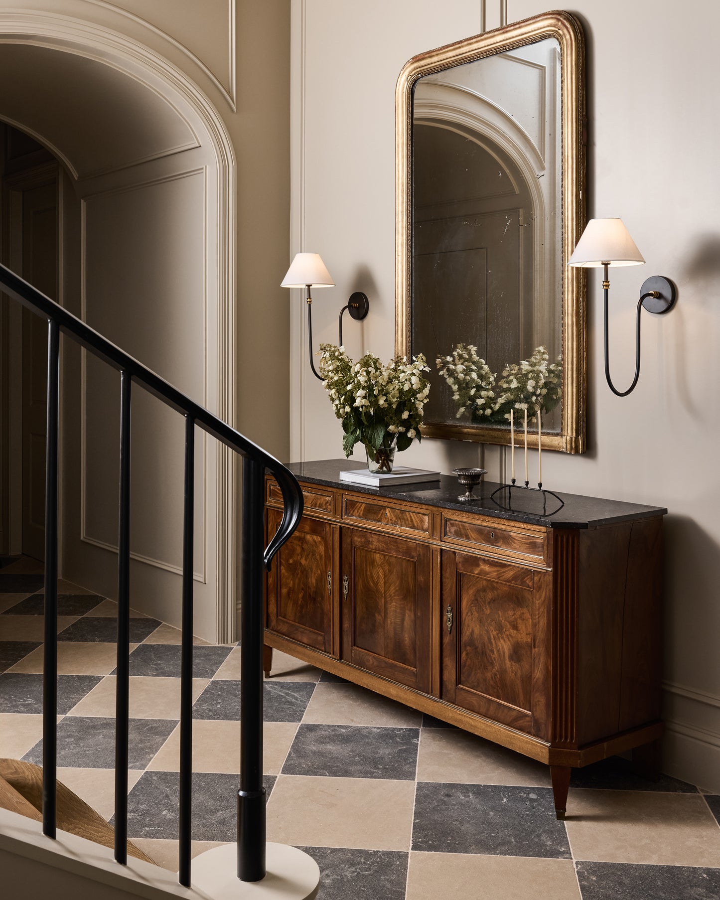 Staircase with wooden sideboard, mirror, checkered limestone flooring, and wall lights in a classic interior setting.